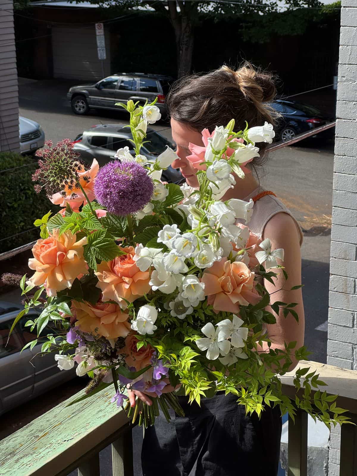 Florist holding an extra large hand-tied bouquet with peach garden roses, white campanula bells and purple allium on a sunny porch, by Kvetka Flower