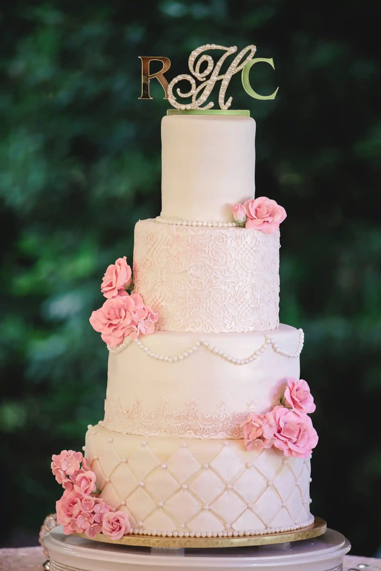 Four-tiered wedding cake with pink flowers and a decorative topper against a blurred green background