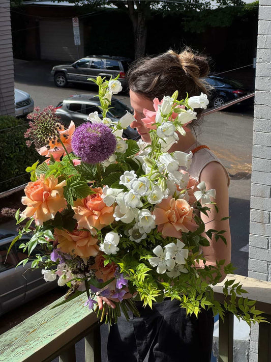 Florist holding an extra large hand-tied bouquet with peach garden roses, white campanula bells and purple allium on a sunny porch, by Kvetka Flower