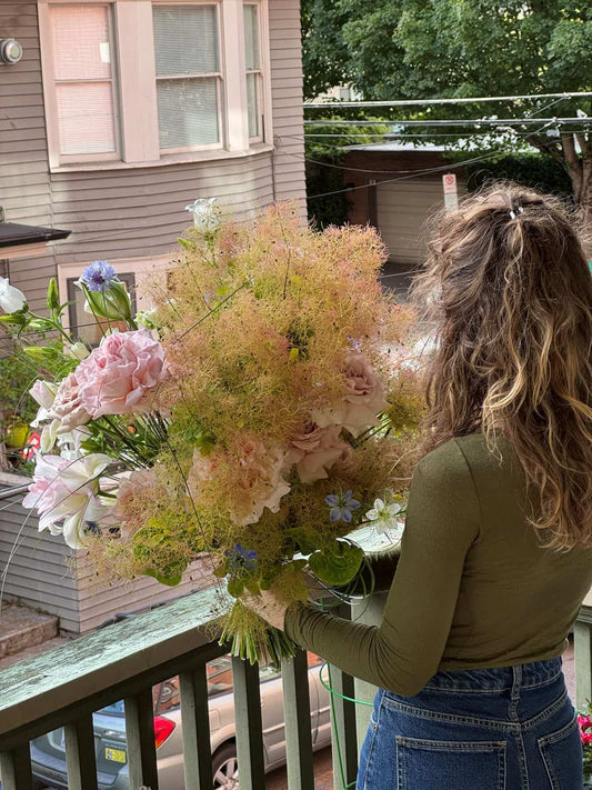 Florist holding a grande hand-tied bouquet with airy smoke bush, pink garden roses, lilies and blue cornflowers on a porch, by Kvetka Flower